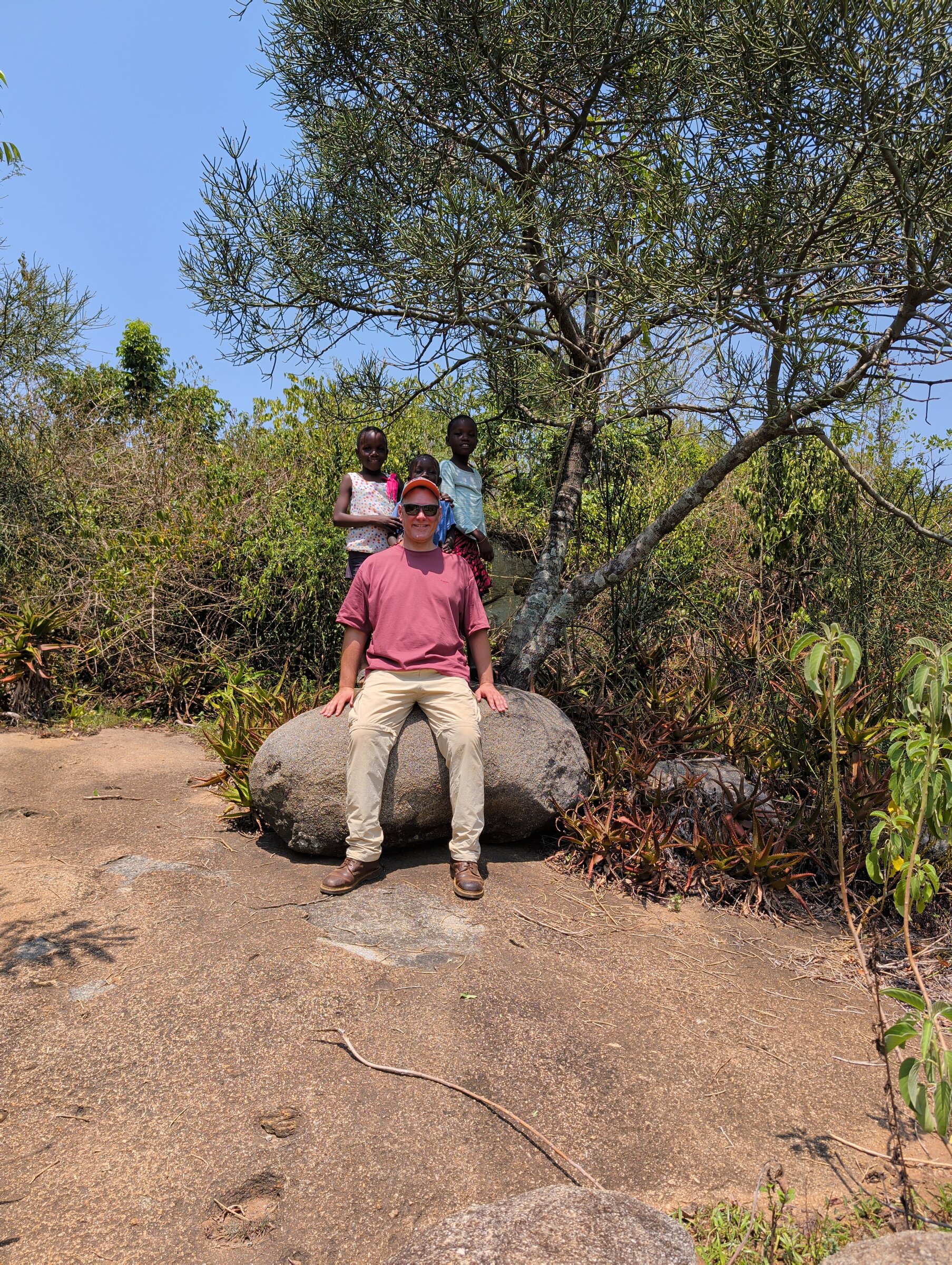 Philipp sitzt auf einem Felsen, hinter ihm stehen drei Kids in der Natur von Siaya